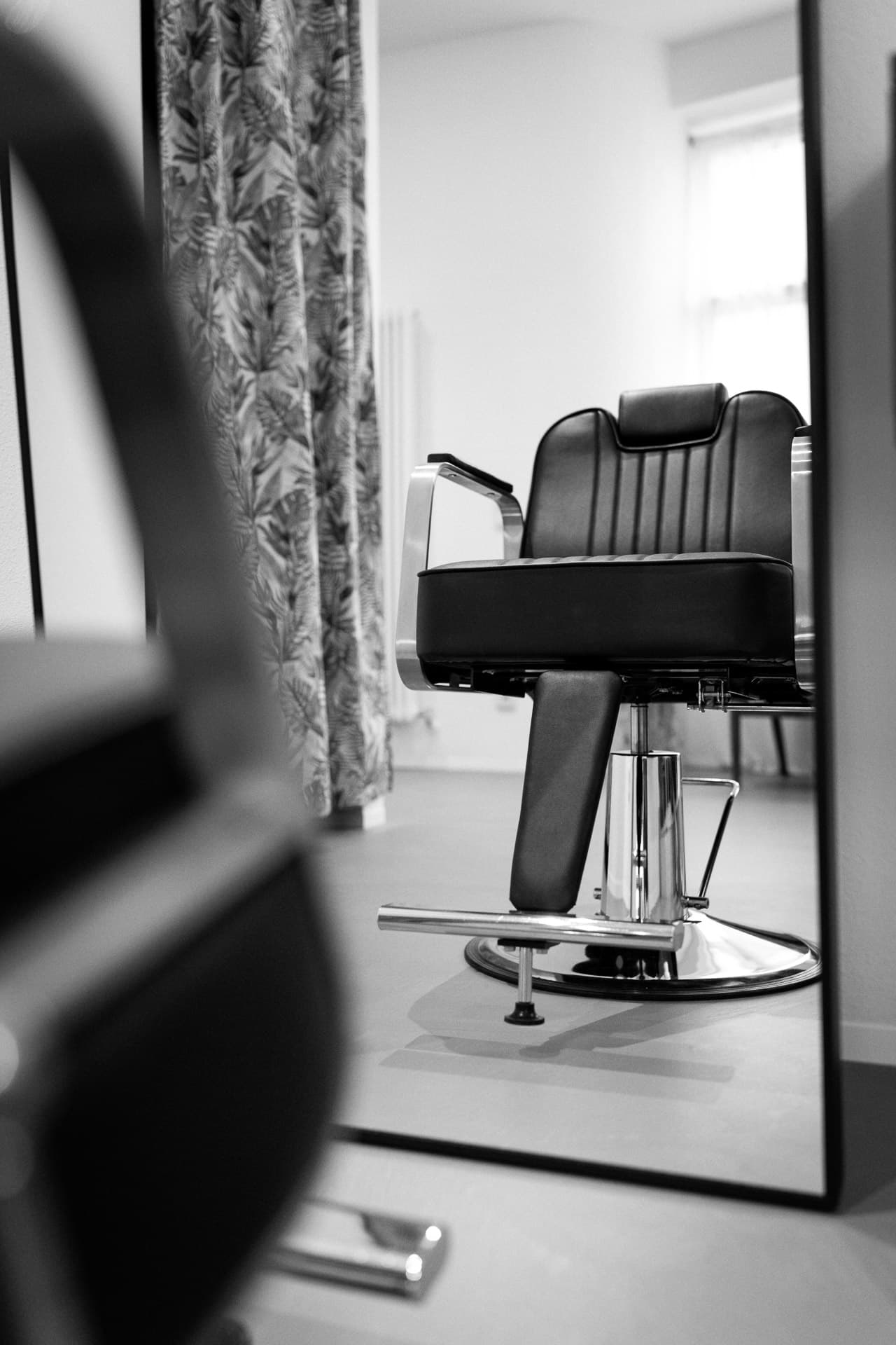 Schwarzweiß: Spiegelung eines schwarzen Friseurstuhls mit Chromfuß und Palmblatt-Vorhang im Salon