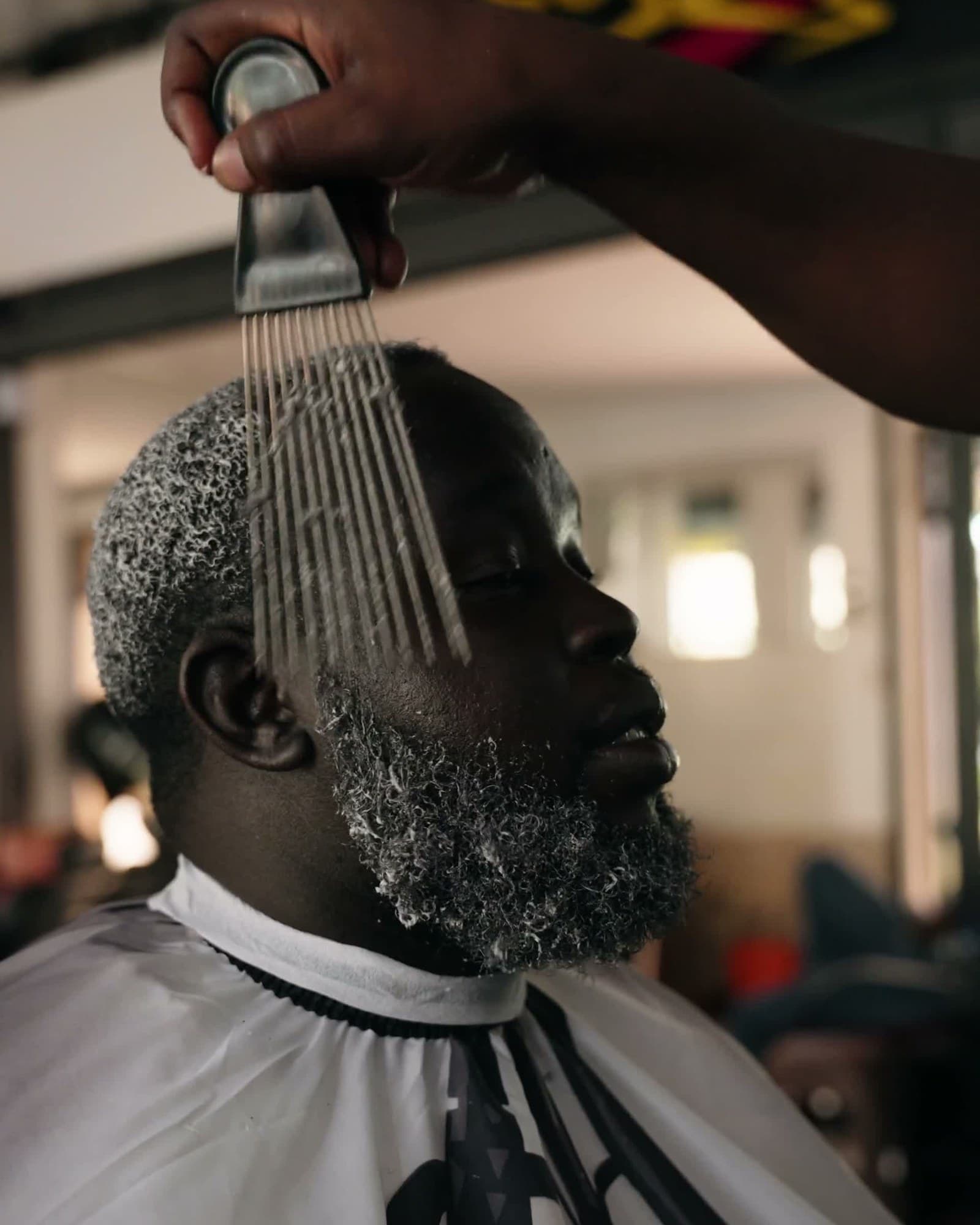 Nahaufnahme: Kunde mit grauem Bart wird mit Afro-Pick frisiert, weißer Barber-Umhang, Jinja