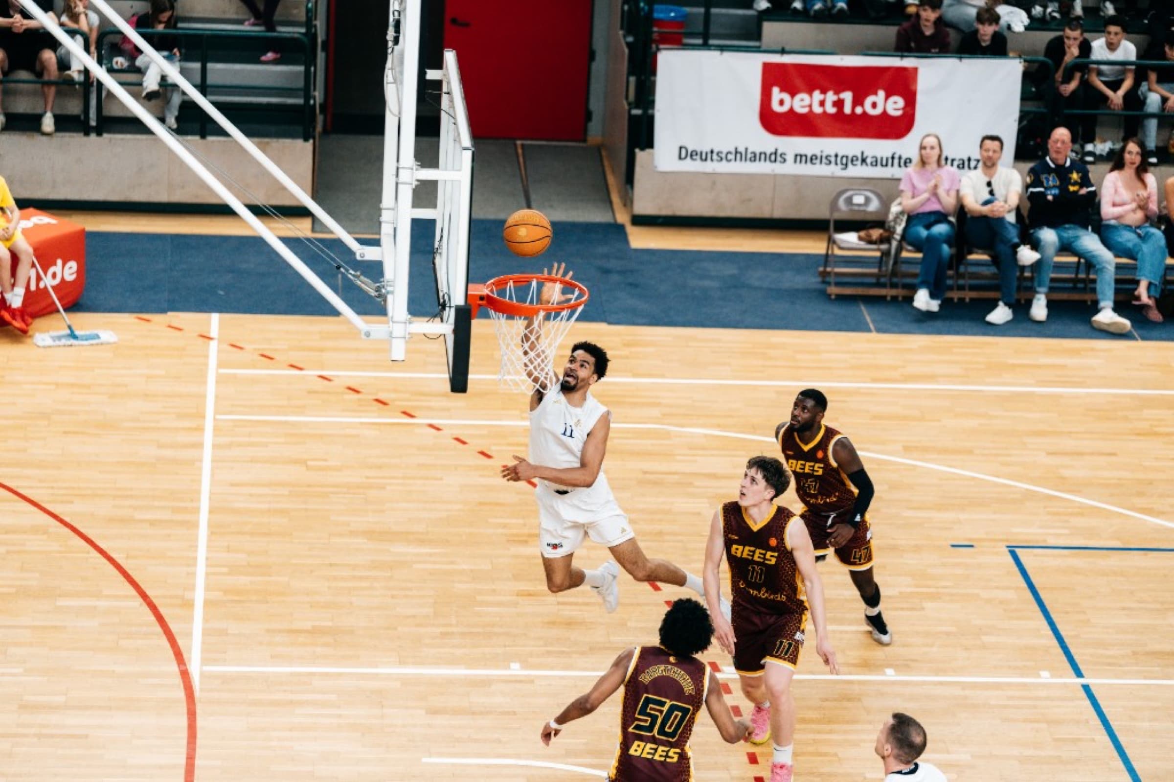 Player in white leaping for a layup over Bees defenders in maroon and gold
