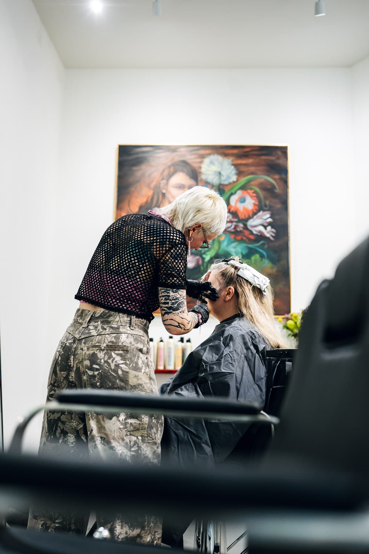 Stylist mit Handschuhen färbt Kundin mit Folien, großes Blumenbild an der Wand im Hintergrund