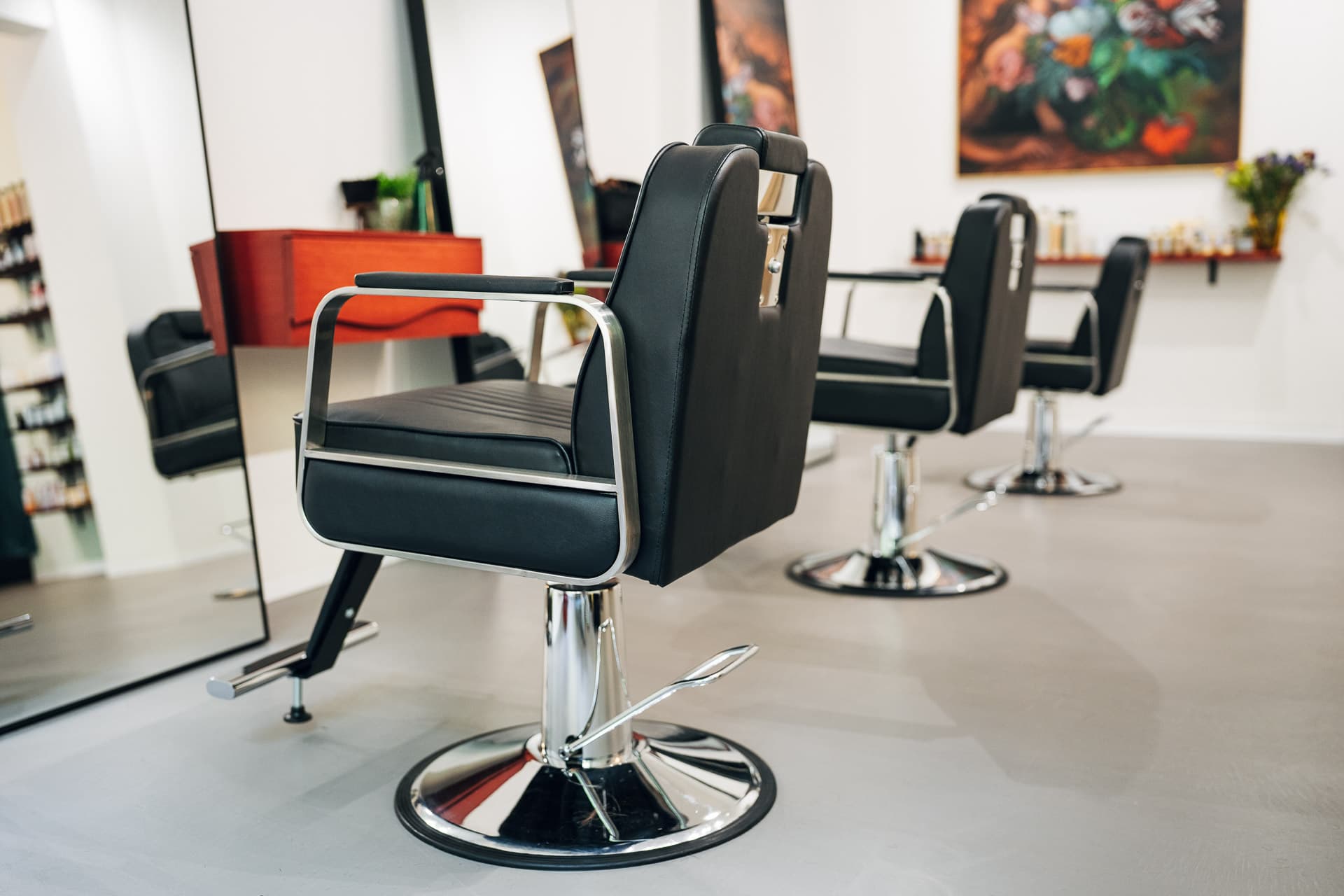 Reihe schwarzer Chrom-Friseurstühle im hellen Salon, Blumenbild und Produktregal hinten