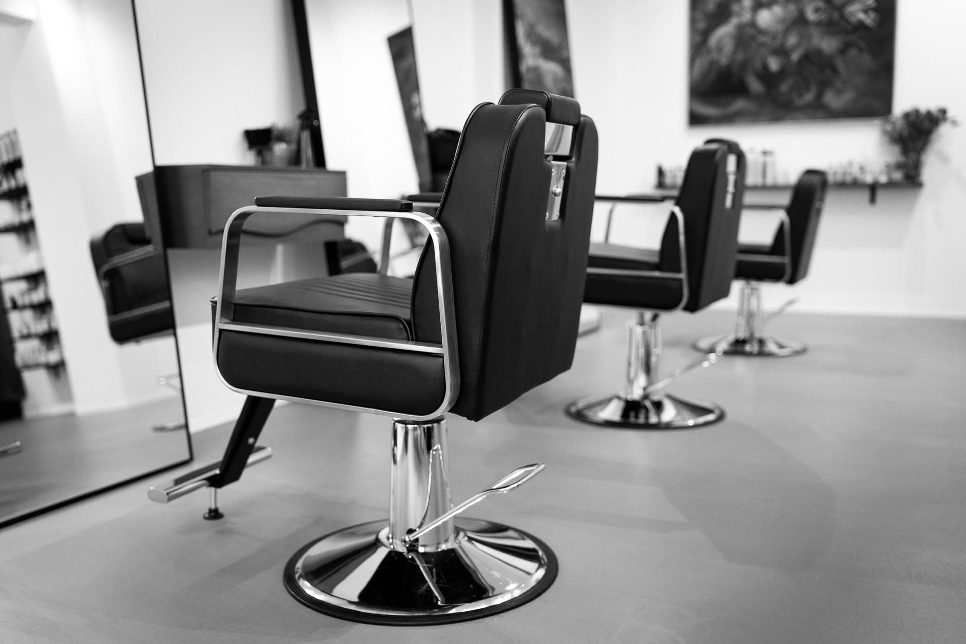 Schwarzweiß: drei Friseurstühle in Reihe mit Chromsockeln und flachem Betonboden