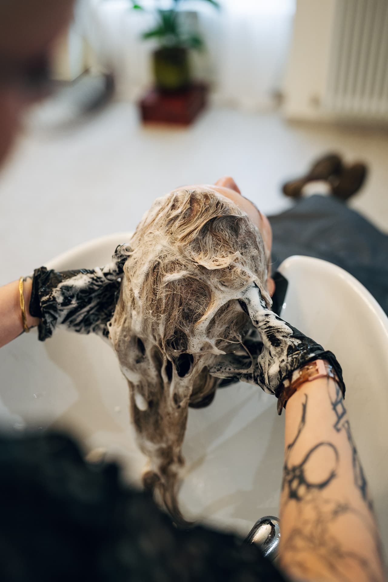 Von oben: Stylist mit Scheren-Tattoo wäscht blondes Haar mit Schaum im weißen Waschbecken
