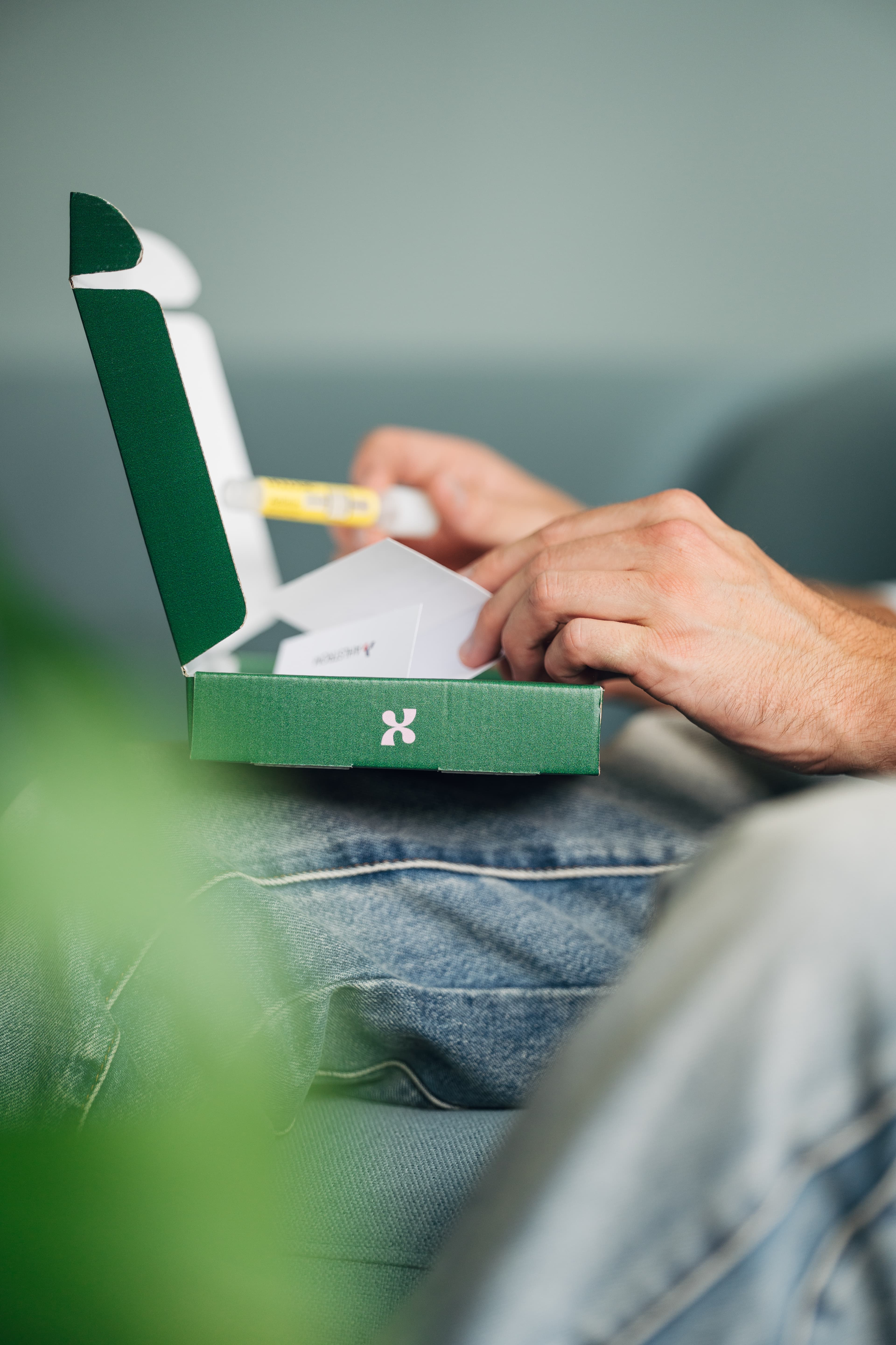 Hände öffnen die dunkelgrüne heymacy-Box auf dem Schoß, gelbes Röhrchen und Karte sichtbar
