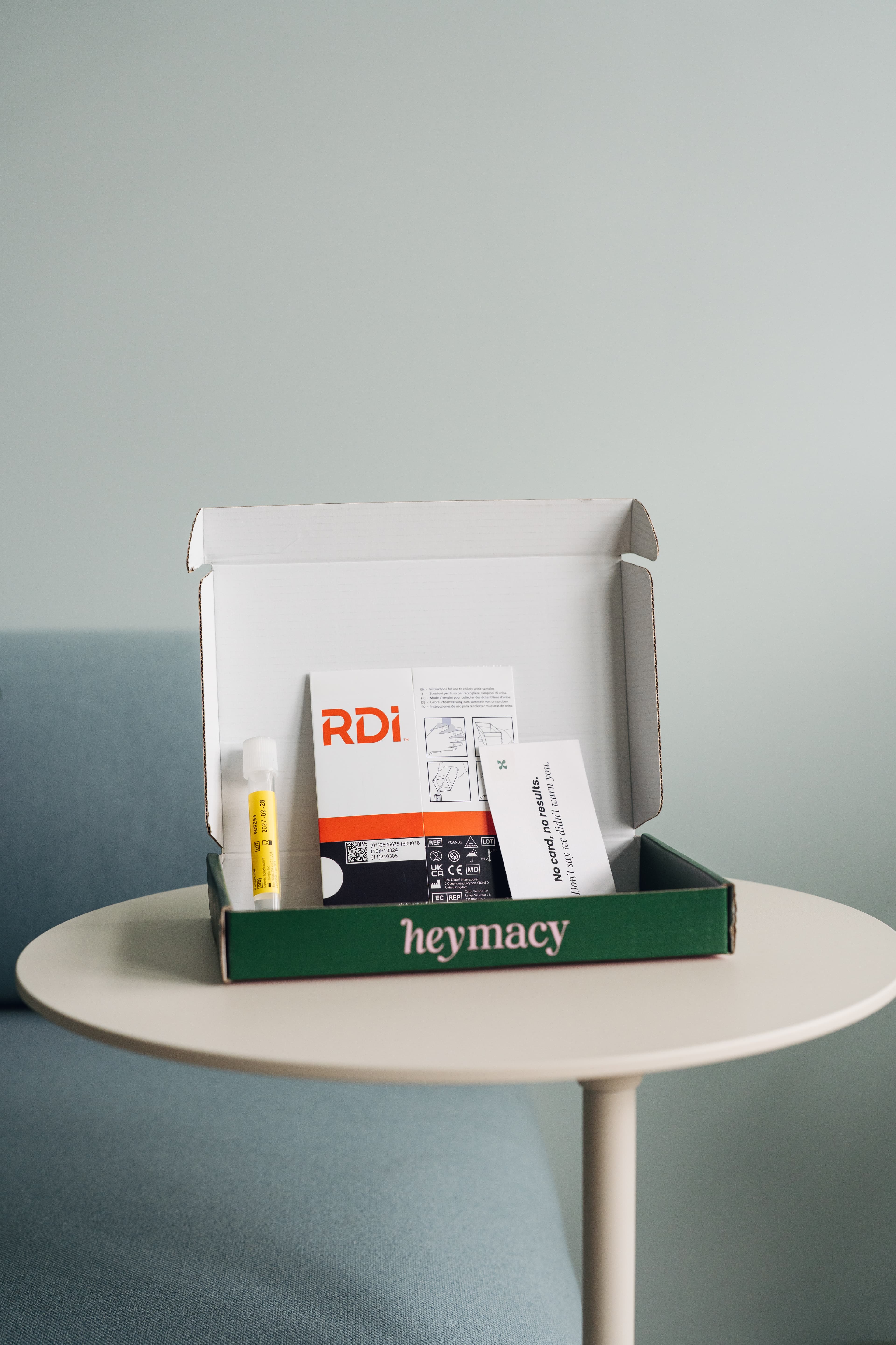 Geöffnete heymacy-Box auf rundem Tisch: gelbes Röhrchen und Anleitung neben Sofa