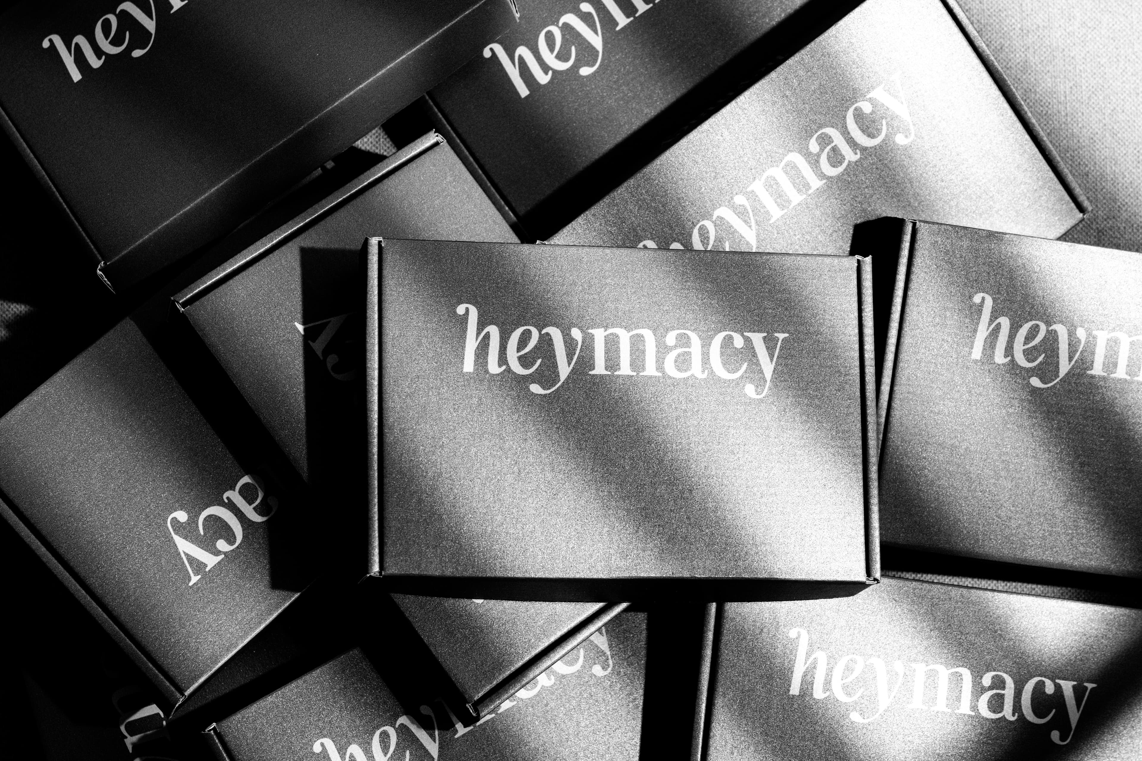 Schwarzweiß: Stapel schwarzer heymacy-Boxen mit weißem Serif-Logo und diagonalen Schatten