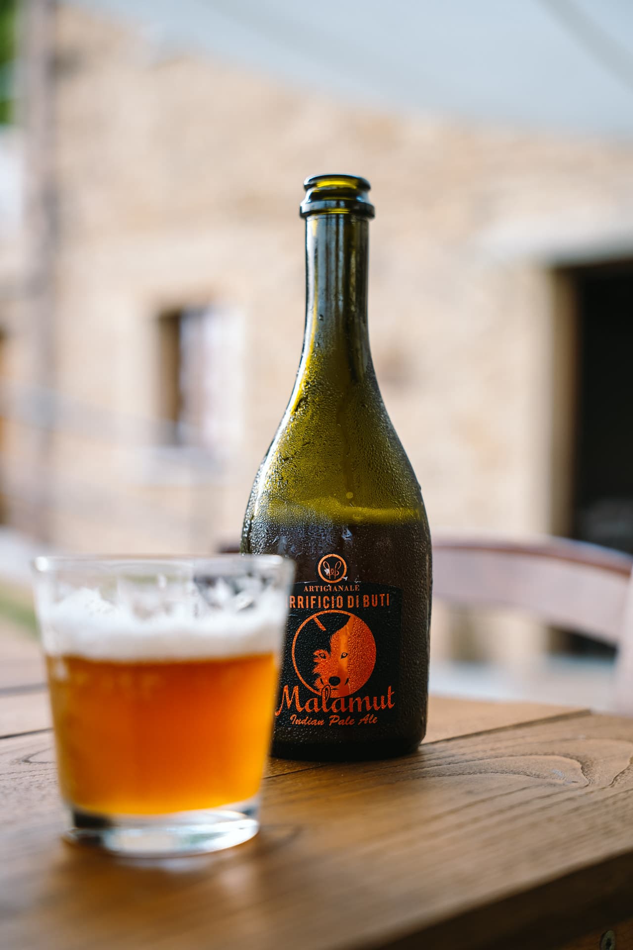 Beschlagene Flasche Malamut IPA und Glas mit Schaum auf Holztisch vor steinerner Fassade