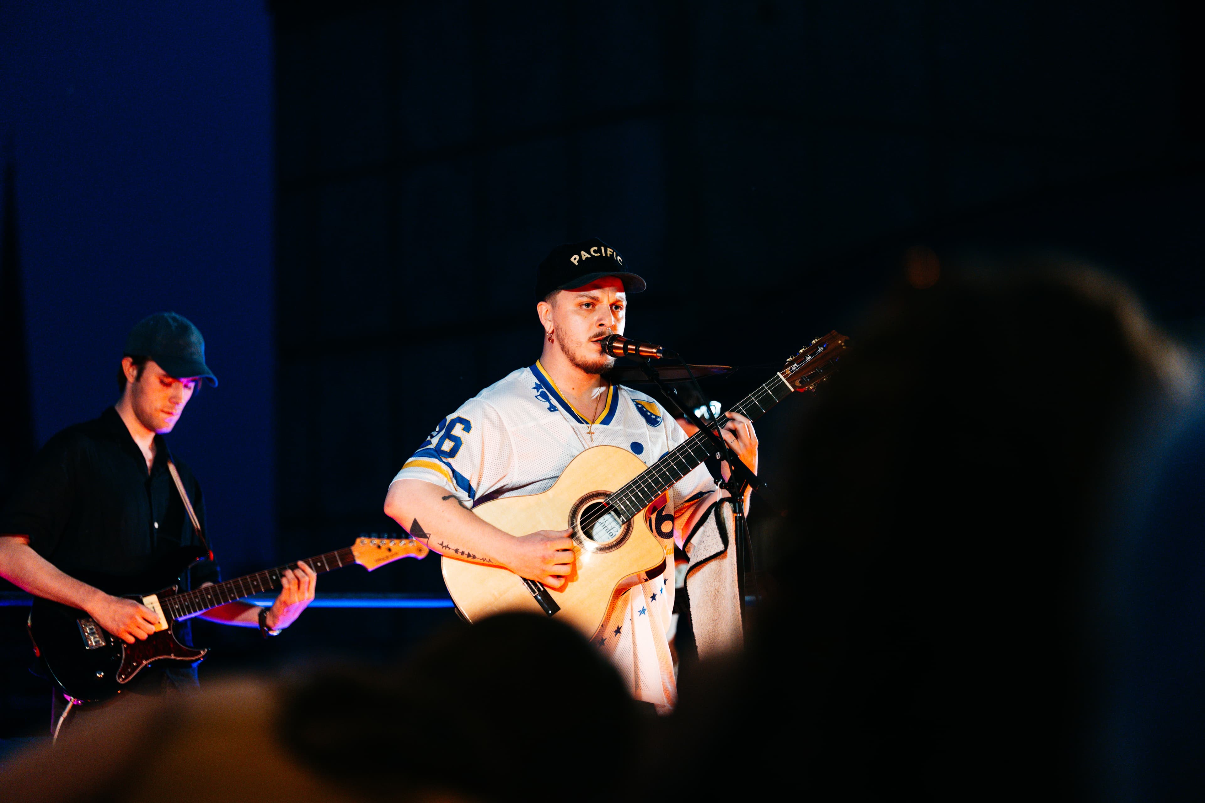 Sänger mit Akustikgitarre und Jersey Nummer 26 auf der Sofar-Bühne, zweiter Gitarrist und Publikum unscharf