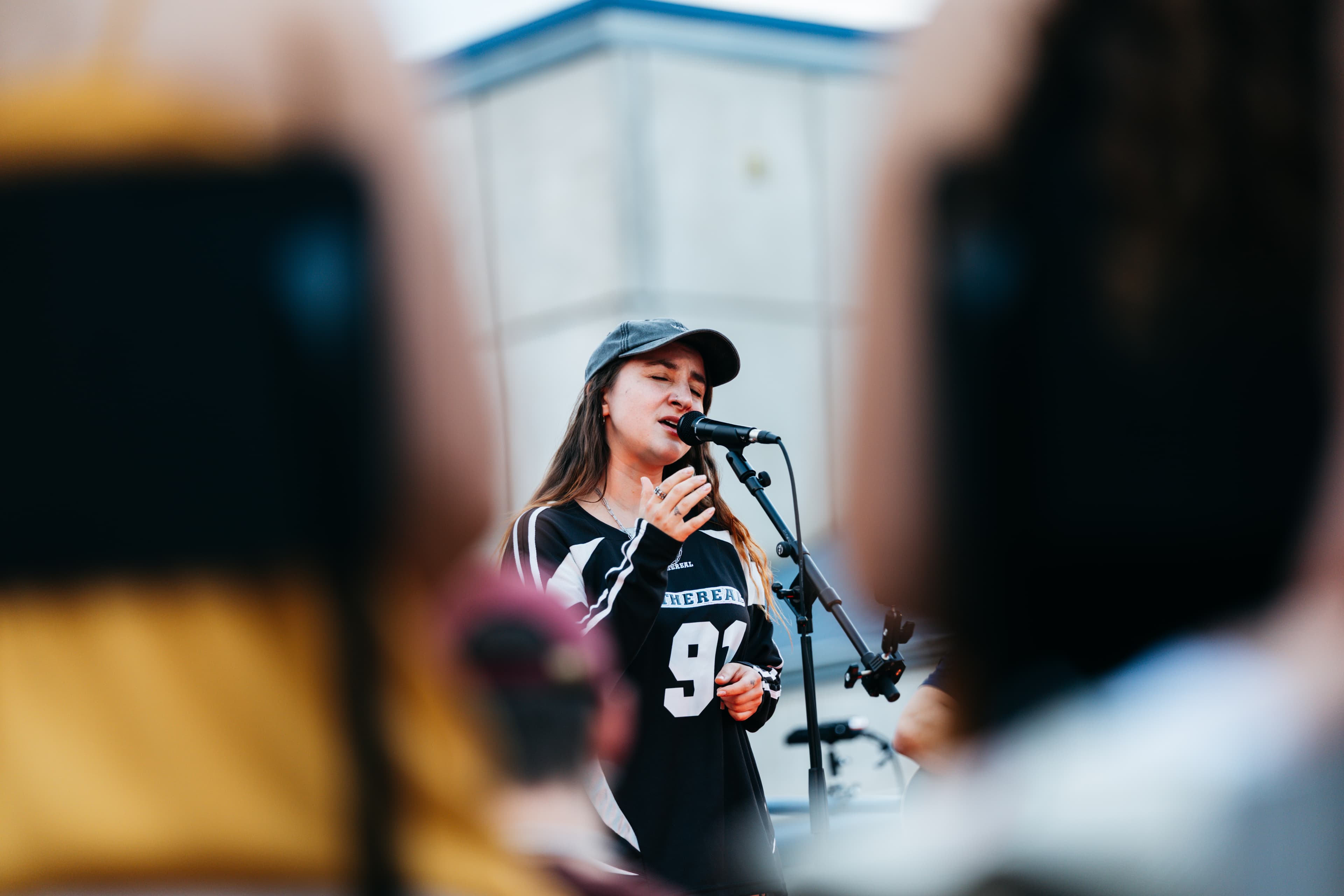 Female singer in a black cap and jersey 91 singing into a mic, audience blurred in the foreground