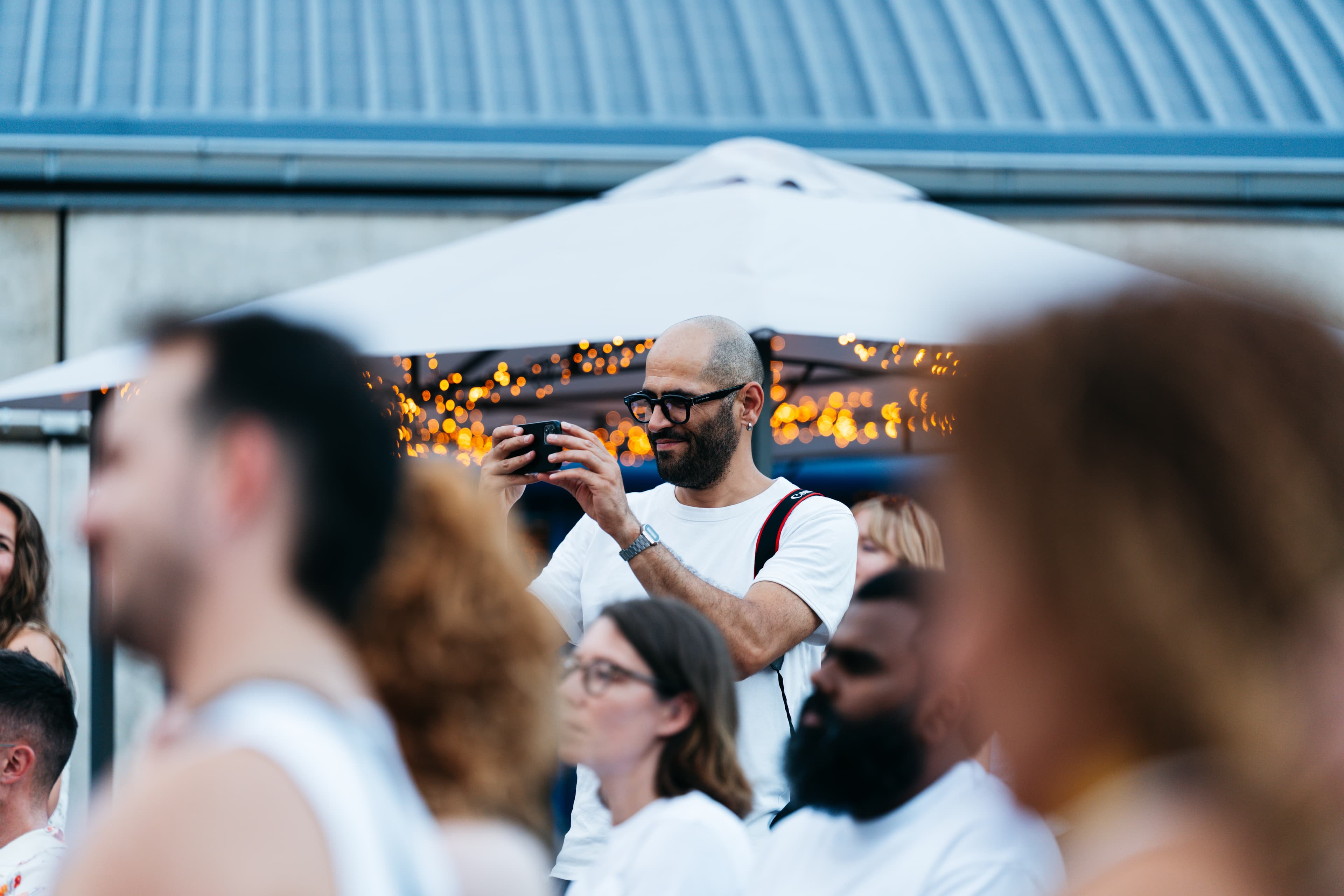 Gast mit Brille und weißem Shirt filmt lächelnd mit dem Smartphone unter Lichterketten