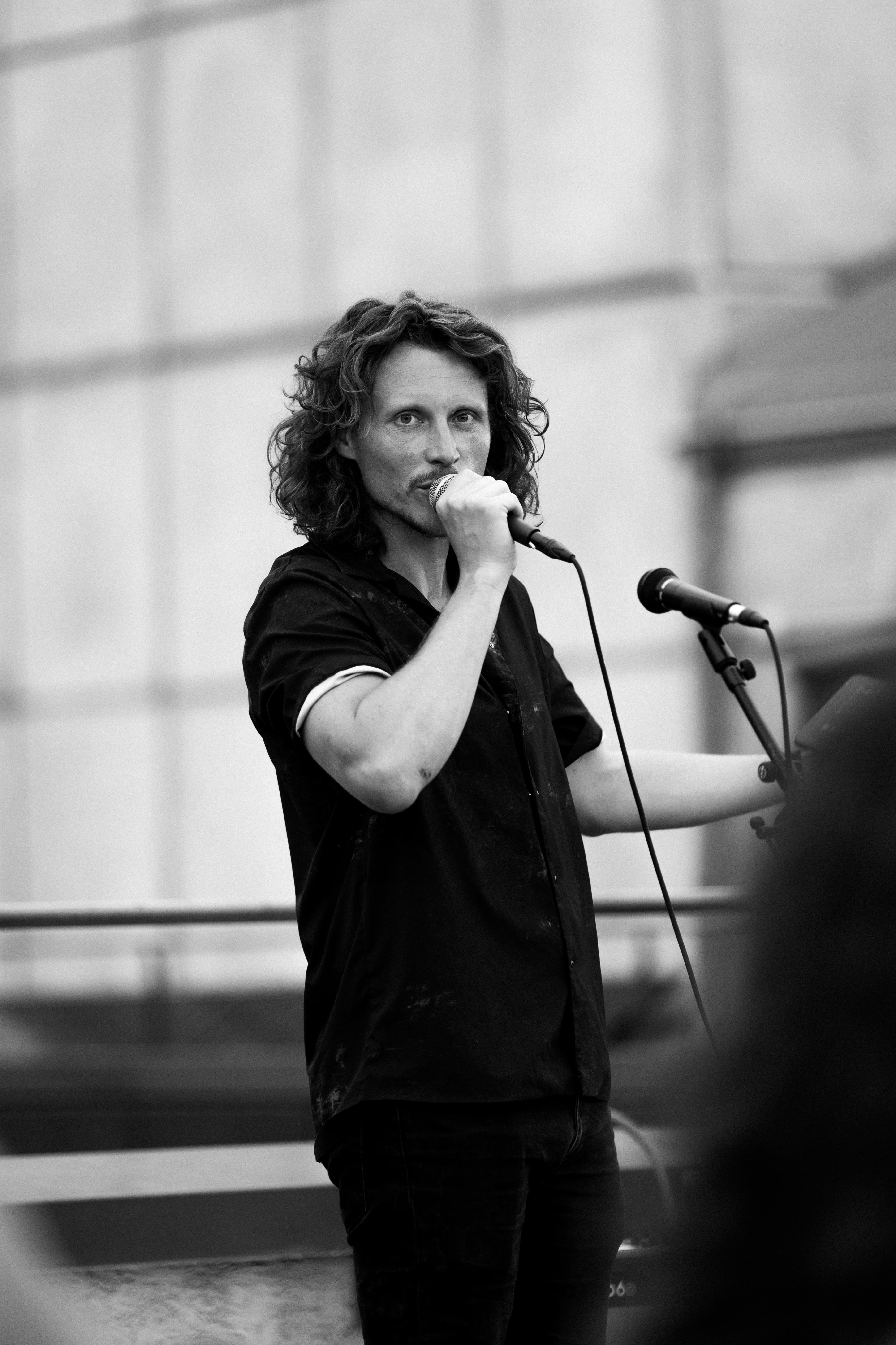Black and white: man with long wavy hair speaking or singing into a handheld mic on the roof