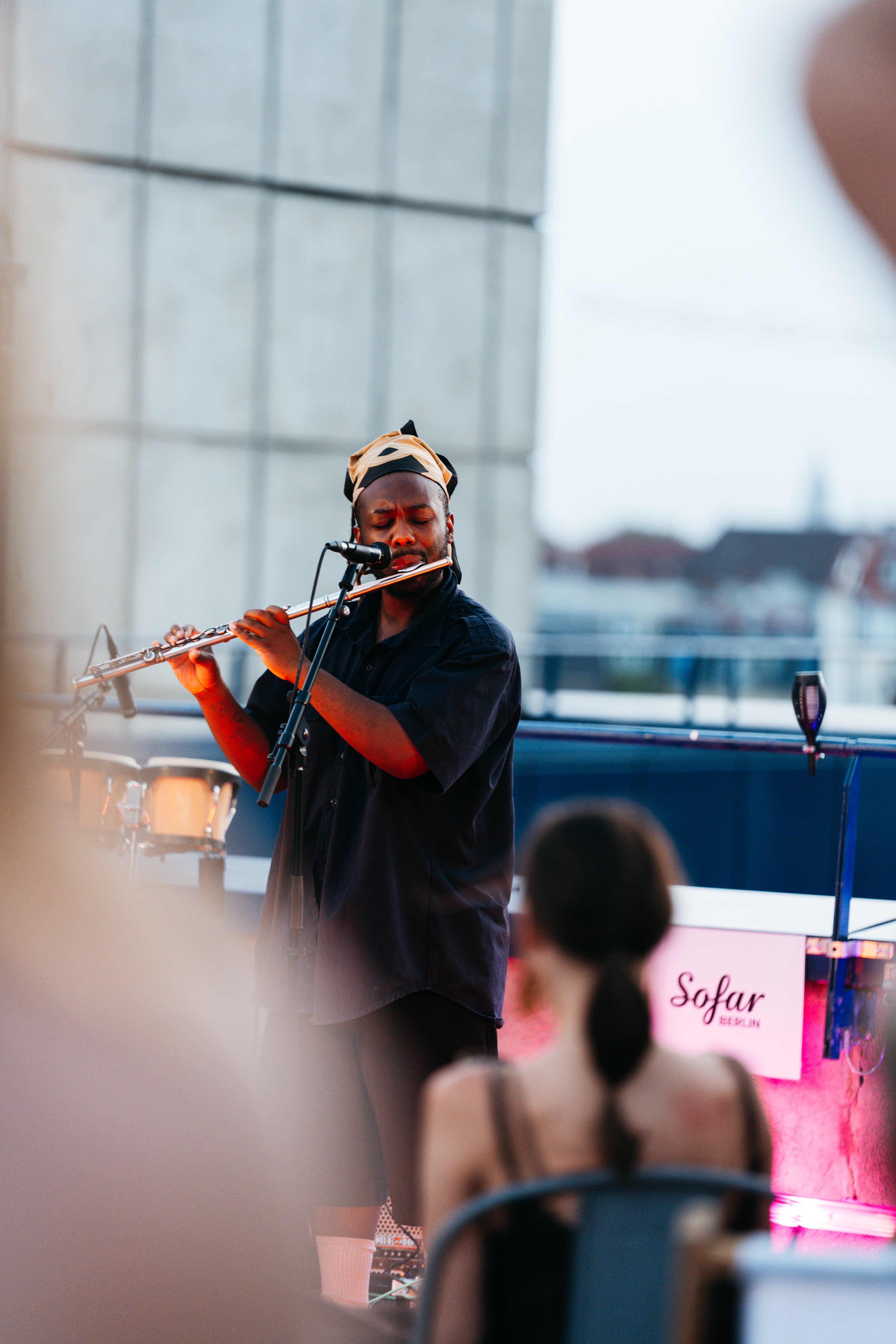 Musician playing flute in a headwrap at Sofar Berlin on the roof, warm stage light, audience blurred