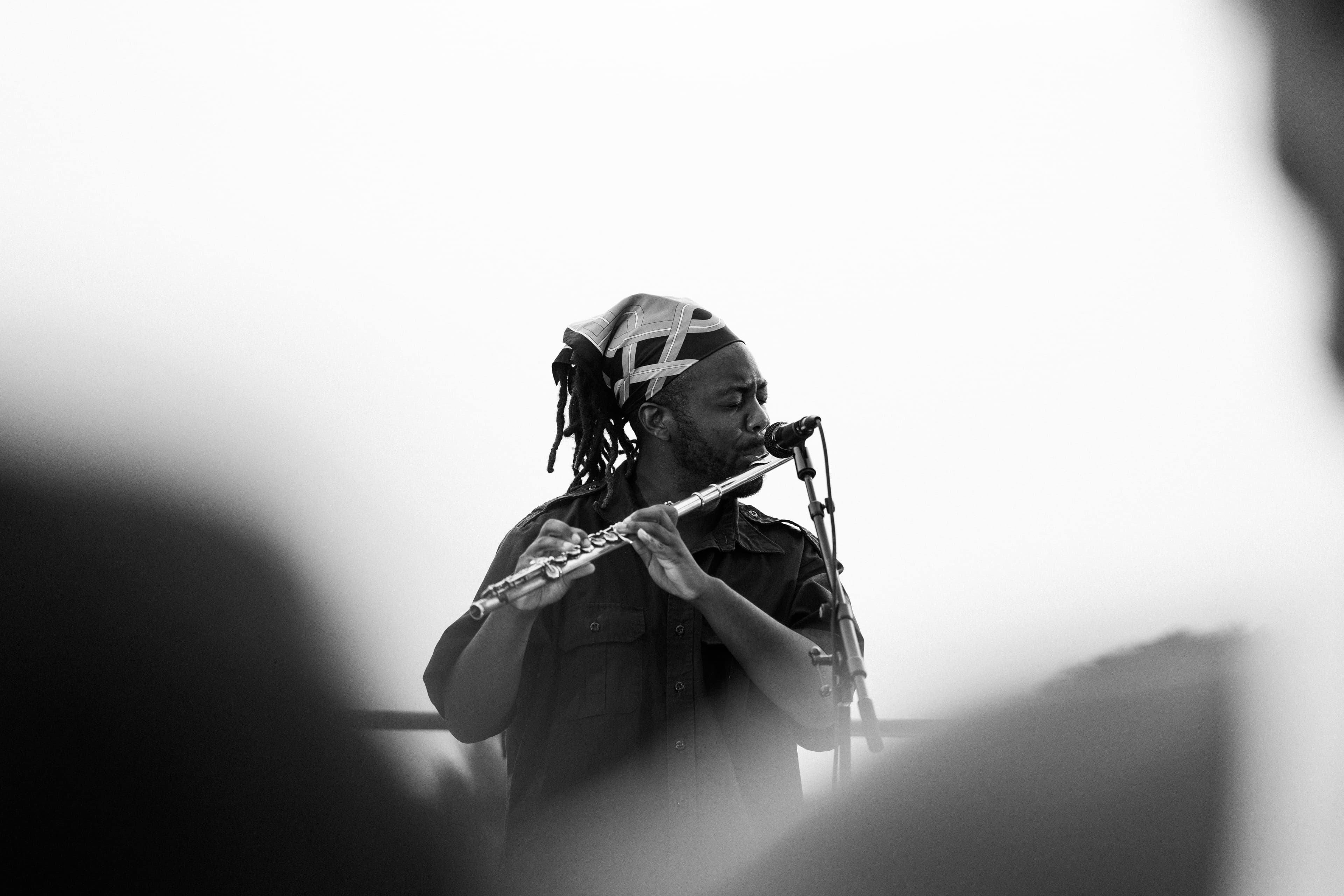 Black and white: flautist with dreadlocks and a patterned headwrap playing into a mic, audience in foreground