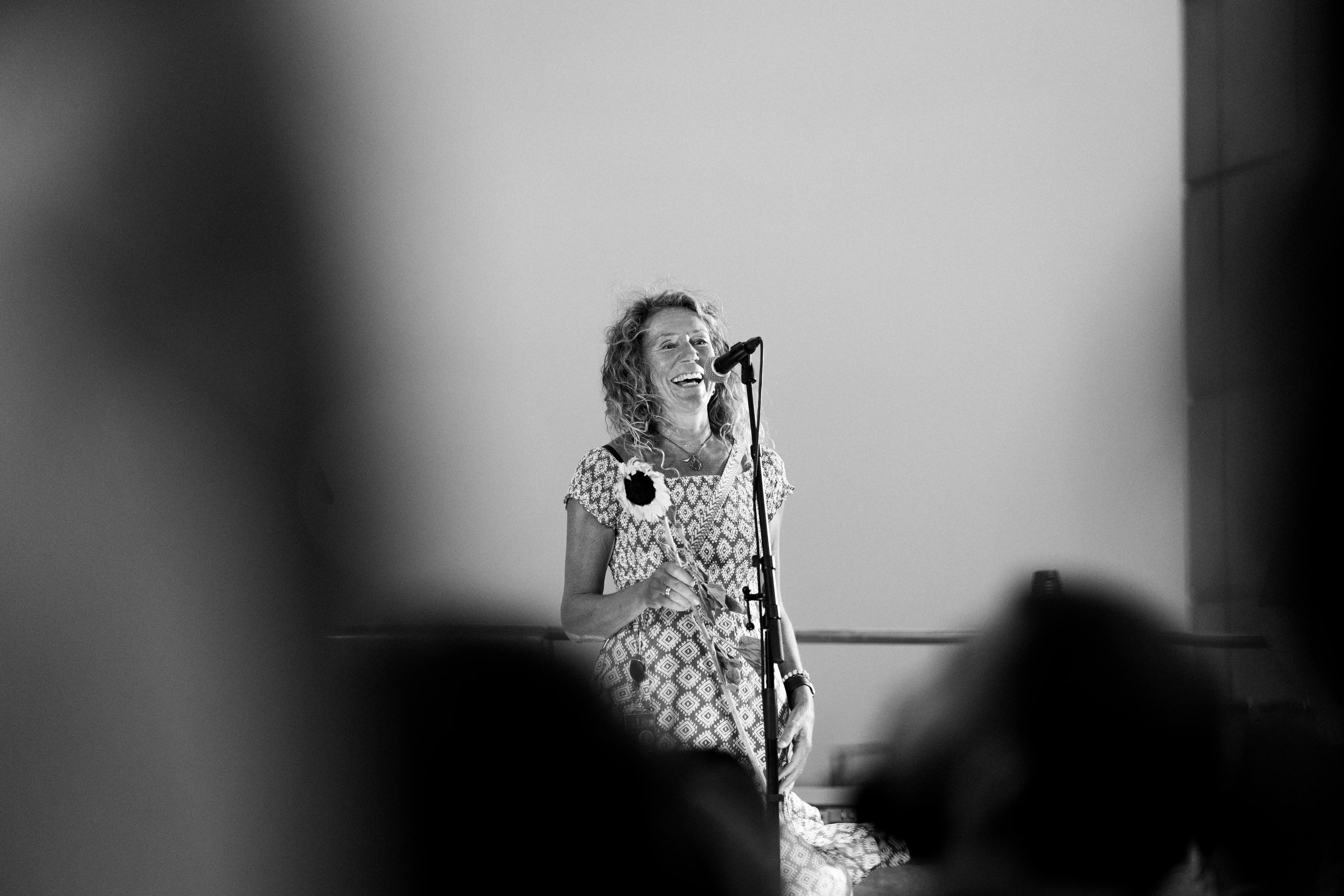 Black and white: smiling woman with curly hair and a sunflower speaking behind a microphone